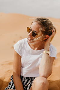 Smiling woman in sunglasses sitting on desert in sunny day