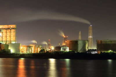 View of illuminated petrochemical plant by river against sky