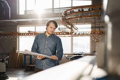 Young inspector holding file while standing by production line at industry