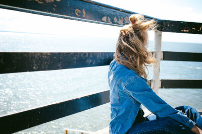 Side view of woman sitting by railing at beach against sky