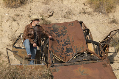 Adult man in cowboy hat with abandoned car in desert, almeria, spain