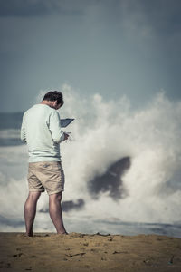 Rear view of man using digital tablet standing against splashing sea waves