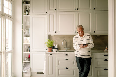 Retired senior man using smart phone while leaning on kitchen counter