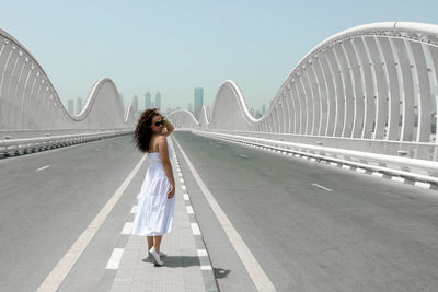 A woman in a white sundress walking on an empty road toward to the city, meydan bridge in dubai