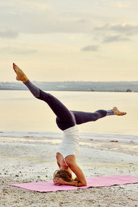 Woman doing headstand at beach against sky during sunset