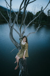 High angle view of young woman sitting on bare tree over lake