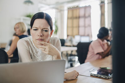 Confused businesswoman looking at laptop while sitting at desk in creative office