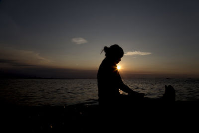 Silhouette woman doing yoga at lakeshore against sky during sunset