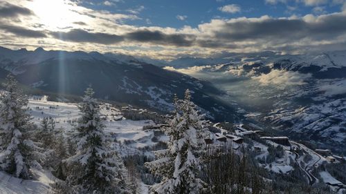 Snow covered trees and mountains against cloudy sky on sunny day