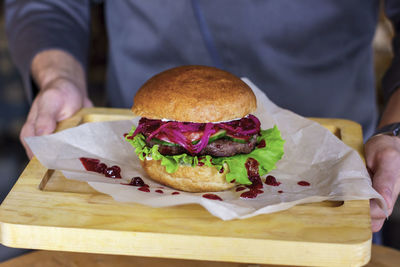 Meat burger with pickled onions, cutlet and cranberry jam on wooden board in man hands