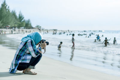 Side view of woman photographing while crouching against people at beach