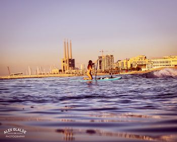 Person on sea by city against sky during sunset