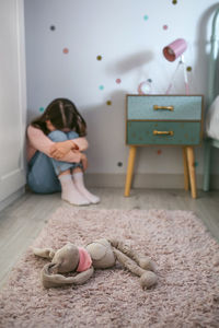 Sad little girl sitting on the floor of her bedroom with stuffed toy lying