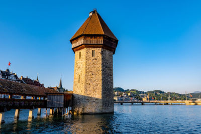 Tower by river and buildings against clear blue sky