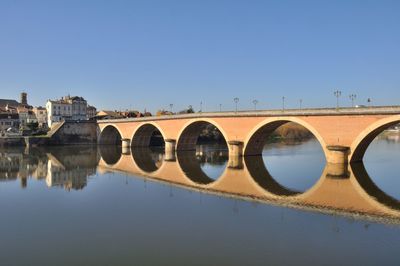 Arch bridge over river by buildings against clear blue sky