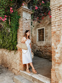 Full length portrait of woman in white summer dress leaning on brick wall.