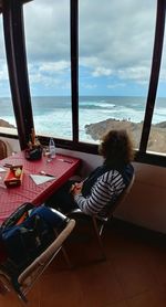 Woman sitting on table by sea against sky seen through window