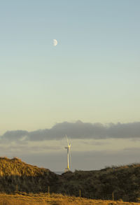 Distance wind turbine on sea with half-moon sky against fields foreground.