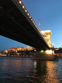 Illuminated suspension bridge over river against sky in city