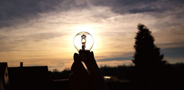 Cropped hand holding light bulb against sky during sunset