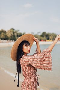 Young woman wearing hat looking away standing at beach against sky