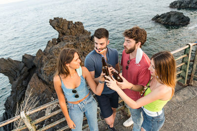 High angle view of friends holding beer standing by railing against sea