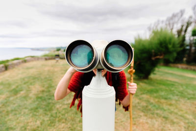 Unrecognizable female hiker with trekking stick looking through binocular telescope while standing on green meadow near sea and exploring nature during pilgrimage through spain
