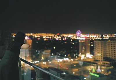 Side view of woman leaning on glass railing against illuminated city at night