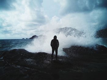 Sea waves splashing on man standing at shore against cloudy sky