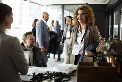 Businesswoman with curly hair standing by disabled entrepreneur while talking to receptionist at registering counter