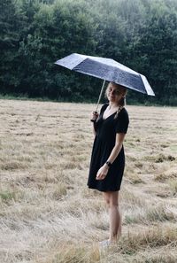 Full length of woman standing on field holding umbrella during rainy day