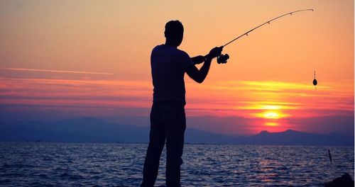 Silhouette man fishing in sea against sky during sunset