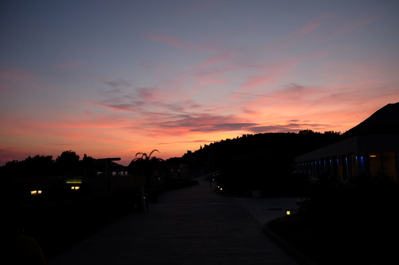 ILLUMINATED FOOTPATH BY SILHOUETTE BUILDINGS AGAINST SKY DURING SUNSET