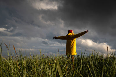 Man in yellow raincoat and red hat with spreading hands wide open standing on field in rainy weather