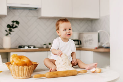 A little boy is sitting on the kitchen table with a rolling pin and a baking dough. mom's assistant