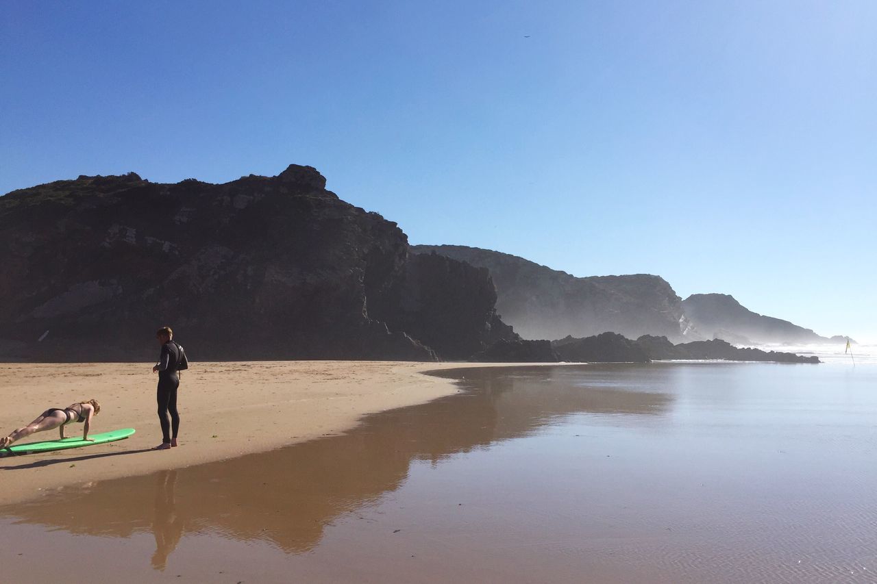 water, full length, sea, clear sky, men, standing, beach, tranquil scene, tranquility, blue, scenics, vacations, solitude, rock formation, beauty in nature, cliff, nature, coastline, outdoors, escapism, shore, remote, calm, in front of, mountain, day