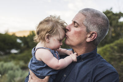 Close-up of father kissing on daughter's forehead while standing against sky in forest