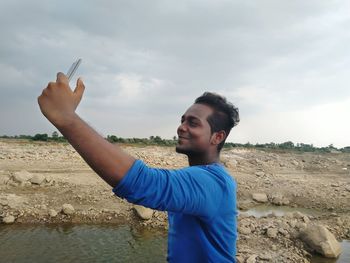 Side view of smiling young man taking selfie through phone on field