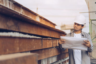 Man holding blueprint while standing at construction site against sky