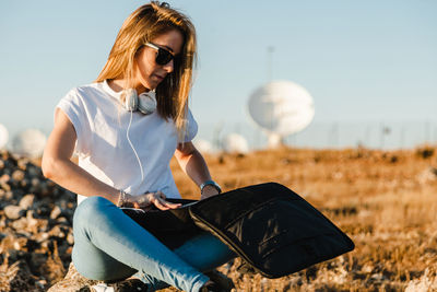 Young woman wearing sunglasses with laptop sitting on land against sky