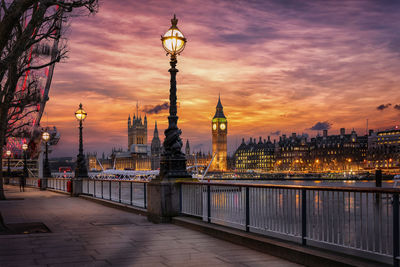 Illuminated buildings by river against sky during sunset