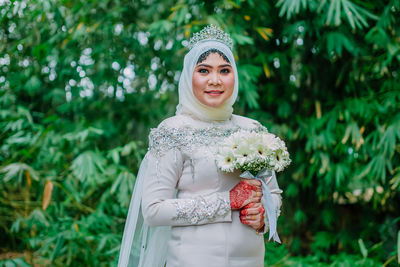 Portrait of smiling bride holding bouquet while standing against trees