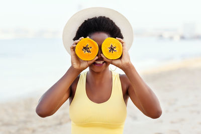 An african girl in a summer straw hat sits on the sand, smiling and closing her eyes with a papaya