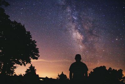 Rear view of man standing by silhouette trees against star field in sky