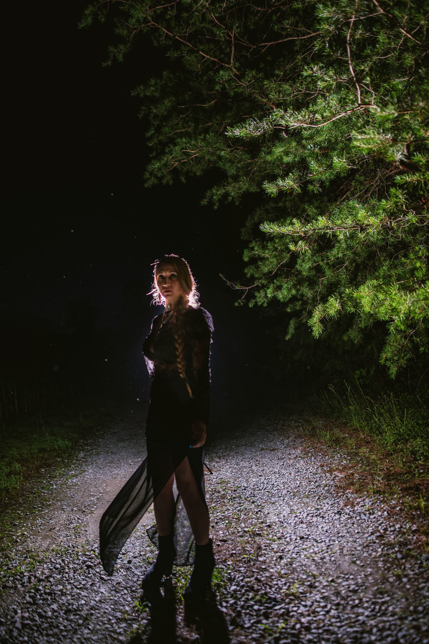 darkness, one person, full length, tree, adult, plant, nature, night, young adult, light, standing, women, forest, outdoors, clothing, footwear, wet, person, lifestyles, looking, fashion, casual clothing