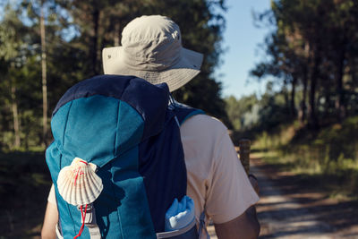 Back view of unrecognizable male traveler with rucksack and wooden stick walking on rough path between trees in summer