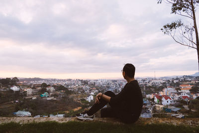 Side view of man sitting on retaining wall over city against sky