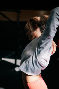Side view of positive young fit female athlete in sportswear performing stretching exercise during workout in dark gym