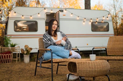 Caucasian woman sits in a wicker chair wrapped in a blanket in the yard near the trailer in autumn