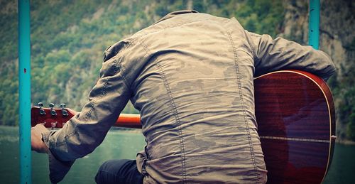 Rear view of man playing guitar while sitting by lake